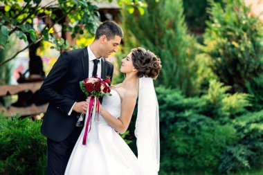 Gelin ve damat bahçede dikiliyor, gelin elinde bir buket kırmızı çiçek tutuyor.