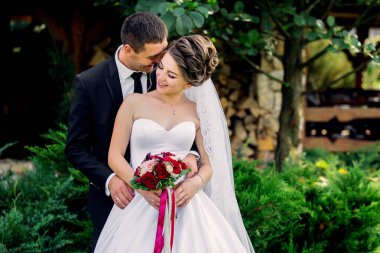 Bir gelin ve damat bahçede fotoğraf çektirmek için poz veriyorlar. Damat takım elbise giyerken gelin beyaz bir elbise ve kırmızı bir buket giyiyor. Sahne mutlu ve romantik.