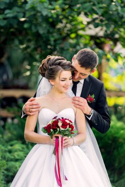 Bir gelin ve damat parkta fotoğraf için poz veriyorlar. Gelin beyaz bir elbise giyiyor ve elinde bir buket kırmızı çiçek tutuyor. Damat siyah takım elbise ve kırmızı kravat takıyor.