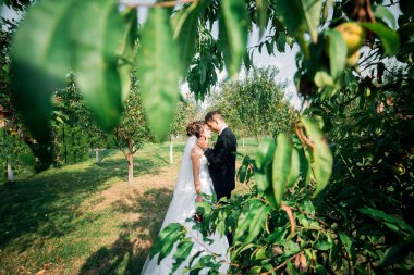 Bir gelin ve damat arka planda bir ağaçla bahçede duruyorlar. Gelin beyaz bir elbise giyiyor ve damat bir takım elbise giyiyor. Birbirlerini kucaklıyorlar.