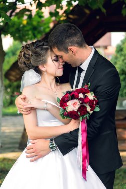 Bir gelin ve damat bir bahçe ortamında birbirlerini kucaklıyorlar. Gelin beyaz bir elbise giyiyor ve damat siyah bir takım elbise giyiyor. Bir buket kırmızı çiçek ve bir saat tutuyorlar.