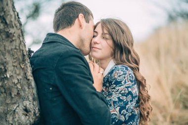 Bir çift, bir ağacın altında kadının boynunu öpen adamla sarılıyor. Sahne romantik ve samimi.