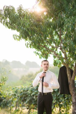 Bir adam ağacın önünde duruyor, takım elbiseli ve kravatlı. Kravatını düzeltiyor ve kameraya bakıyor. Sahne resmi ya da profesyonel bir ortam öneriyor.