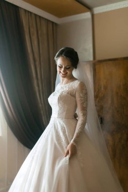 Gelinlikli bir kadın perdenin önünde duruyor. Gülümsüyor ve kameraya bakıyor.