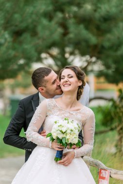 Bir gelin ve damat birbirlerine sarılıp öpüşüyorlar. Gelin elinde bir buket çiçek tutuyor.