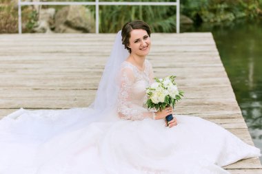 Kucağında bir buket çiçekle bir gelin rıhtımda oturuyor. Beyaz bir elbise ve duvak takıyor.