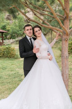 Bir gelin ve damat bir ağacın önünde fotoğraf için poz veriyorlar. Gelin beyaz bir elbise giyiyor ve damat bir takım elbise giyiyor. Sahne mutlu ve romantik.