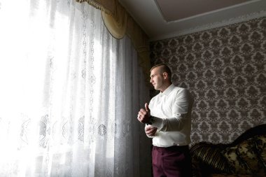 Beyaz gömlekli bir adam pencerenin önünde duruyor, kravatını düzeltiyor. Odada antika bir his var, arka planda bir kanepe ve masada bir vazo. Adamın acelesi var.