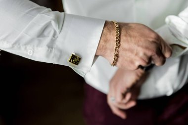 Bir adam takım elbise ve kravat giymeye hazırlanıyor. Altın bir bilezik ve kol düğmesi takıyor.