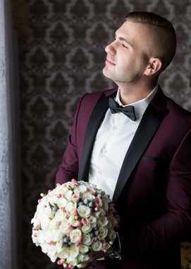 Elinde bir buket çiçek tutan takım elbiseli bir adam. Adam siyah bir kravat ve kırmızı bir ceket giyiyor. Çiçekler beyaz ve pembe. Adam pencereden dışarı bakıyor.