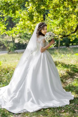 Beyaz elbiseli bir kadın elinde bir buket çiçek tutuyor. Bir duvak takıyor ve bir tarlada duruyor.