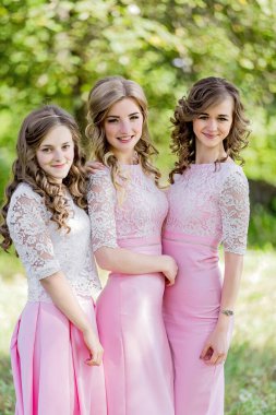 Üç genç kadın pembe elbiseler giyip bir tarlada duruyorlar ve gülümsüyorlar. Sahne parlak ve neşeli, kızlar birlikte poz veriyorlar.