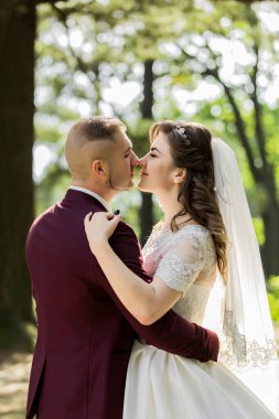 Bir gelin ve damat ormanda birbirlerine sarılıyorlar. Gelin beyaz bir elbise giyiyor ve damat kırmızı bir takım elbise giyiyor. Sahne mutlu ve romantik.