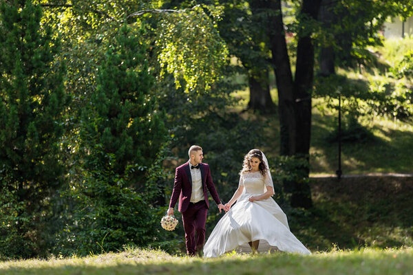 A bride and groom are walking through a forest, holding hands. The bride is wearing a white dress and the groom is wearing a red suit. The scene is peaceful and romantic