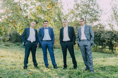 Takım elbiseli ve kravatlı dört adam çimen tarlasında fotoğraf çektirmek için poz veriyor. Sahne resmi ve rahat, çünkü erkekler iş kıyafeti giymiş ama daha rahat bir ortamda duruyorlar.