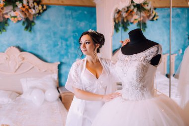 Gelin, yatak odasında gelinliğin önünde dikiliyor. Elbise beyaz ve dantel bir yakası var. Gelin bir taç ve beyaz bir bornoz giyiyor. Odada mavi bir duvar ve bir ayna var.