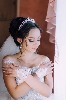 Taçlı ve beyaz elbiseli bir kadın kollarına sarılıyor. Mutlu ve kendinden emin görünüyor.