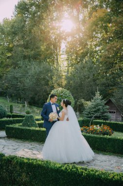 Bir gelin ve damat, çalılar ve çiçeklerle çevrili bir bahçede duruyorlar. Gelin beyaz bir elbise giyiyor ve damat mavi bir takım elbise giyiyor. İkisi de gülümsüyor ve kameraya bakıyor.