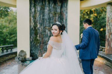 Bir gelin ve damat bahçede birlikte yürüyorlar. Damat mavi bir takım giyerken gelin beyaz bir elbise ve taç giyiyor. Sahne romantik ve samimi.