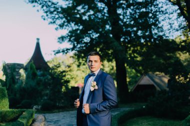 Mavi takım elbiseli ve kravatlı bir adam yakasında çiçekle bir binanın önünde duruyor.