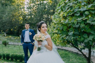 Gelin ve damat parkta, gelin elinde buketle dikiliyorlar. Sahne romantik ve mutlu.