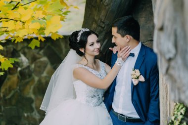 Gelin ve damat bir ağacın yanında, gelin beyaz bir elbise ve damat mavi bir takım elbise giyiyor.