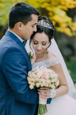 Bir gelin ve damat bir buket çiçek tutarken birbirlerine sarılıyorlar. Gelin beyaz bir elbise giyiyor ve damat mavi bir takım elbise giyiyor. Sahne bir düğün fotoğrafı. Aşkın görüntüsü.