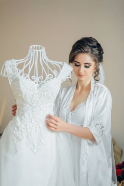 Bir kadın beyaz bir elbisenin önünde duruyor, onu incelemek için tutuyor. Elbise bir mankenin üzerinde ve kadın da müstakbel gelin.