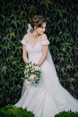 Beyaz elbiseli bir kadın yeşil bir duvarın önünde duruyor. Elinde bir buket çiçek ve duvak tutuyor.