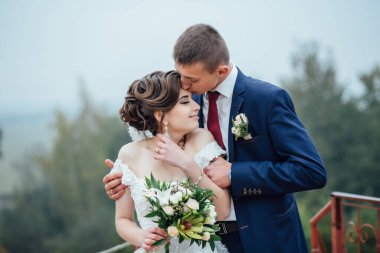 Bir gelin ve damat birbirlerine sarılıyorlar ve gelin bir buket tutuyor. Sahne mutlu ve romantik.