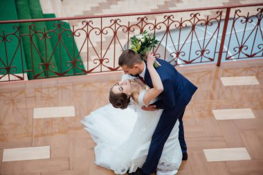 Bir gelin ve damat balkonda birbirlerine sarılıyorlar. Gelin beyaz bir elbise giyiyor ve damat mavi bir takım elbise giyiyor. Sahne romantik ve mutlu.