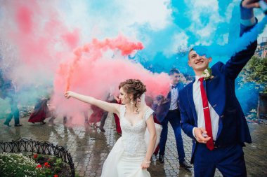 Gelin ve damadın etrafı bir grup insanla çevrili ve gelin elinde kırmızı ve mavi bir duman bombası tutuyor. Sahne kutlama ve neşe dolu, çünkü çift arkadaşlarıyla çevrili.