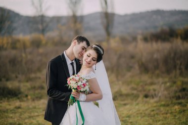 Gelin ve damat bir tarlada duruyorlar, gelin bir buket çiçek tutuyor. Çiftler düğünlerini kutlarken sahne romantik ve mutlu.