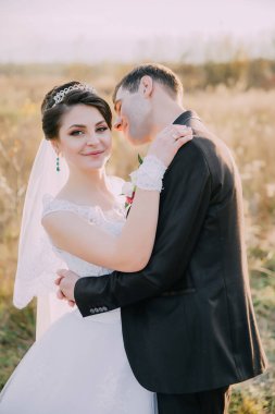 Bir gelin ve damat tarlada sarılıyor. Gelin beyaz bir elbise giyiyor ve damat siyah bir takım elbise giyiyor. Sahne mutlu ve romantik.