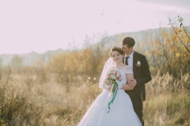 Bir gelin ve damat, arka planda güzel bir dağ manzarası olan bir tarlada duruyorlar.