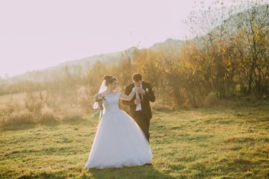 Gelin ve damat, elinde buketle bir tarlada yürüyorlar. Sahne mutlu ve romantik.