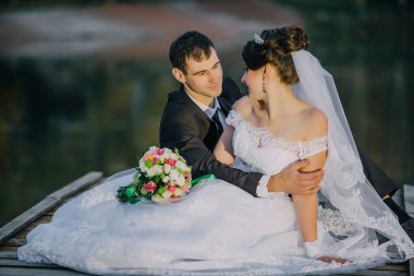 Gelin ve damat rıhtımda oturuyor ve gelin elinde buket tutuyor. Sahne romantik ve samimi.