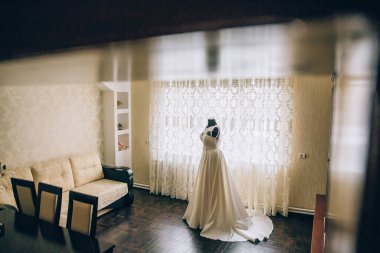 Bir gelin oturma odasında düğününe hazırlanıyor. Oda bir kanepe ve sandalyelerle süslenmiş ve perdeli bir pencere var. Gelin perdenin önünde duruyor.