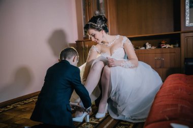 Bir gelin ve damat düğünleri için hazırlanıyorlar. Damat ayakkabılarını giymesine yardım ederken gelin yerde oturuyor.
