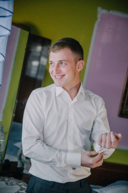 Beyaz gömlekli bir adam pembe bir duvarın önünde duruyor. Gülümsüyor ve gömleğini düzeltiyor.