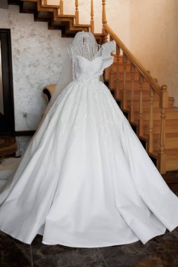 Beyaz bir gelinlik, merdivenli bir odadaki bir mankenin üzerinde sergileniyor. Elbise uzun ve zarif, üzerinde bir peçe var. Sahne bir kutlama ve romantizmdir.