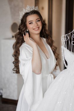 Beyaz elbiseli bir kadın, üzerinde beyaz bir elbise asılı bir şifoniyerin önünde duruyor.