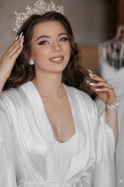 Beyaz bornoz ve taç giyen bir kadın gülümsüyor. Kolye takıyor ve elleri başının üstünde.