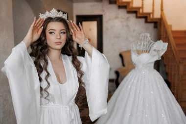 Kafasında taç olan beyaz elbiseli bir kadın şifoniyerin önünde duruyor.