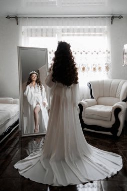 Beyaz elbiseli bir kadın aynanın önünde duruyor ve düğüne hazırlanıyor. Oda bir sandalye ve bir pencere ile süslenmiş, ve masanın üzerinde bir vazo var.