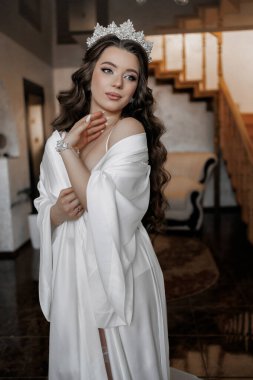 Beyaz elbiseli bir kadın taç takıyor ve ellerini yüzüne dayıyor. Arka planda merdiven olan bir odada duruyor.