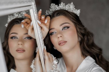 Bir kadın aynanın önünde duruyor, taç takıyor ve beyaz bir elbise giyiyor. Elinde bir kolye tutuyor.