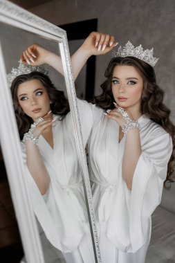 Taçlı ve beyaz cüppeli bir kadın aynanın önünde duruyor. Bir resim için poz veriyor ve kendinden emin ve zarif.