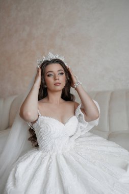 Beyaz elbiseli bir kadın kanepede oturuyor ve taç takıyor. Zarif ve özgüvenli görünüyor.