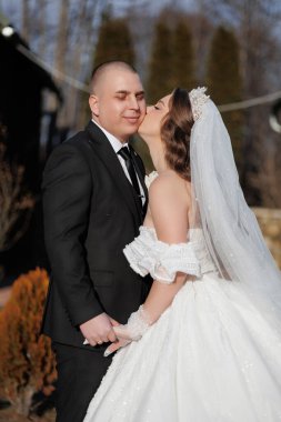 Bir gelin ve damat birbirlerinin yanaklarını öpüyor. Gelin beyaz bir elbise giyiyor ve damat siyah bir takım giyiyor.